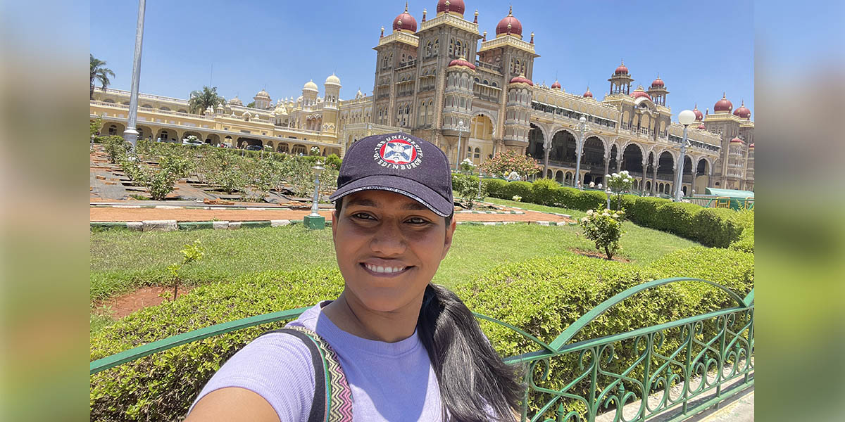 Meera standing in front of a grand building on a sunny day. She is wearing a University of Edinburgh cap.