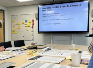Content Design Monthly meet-up session. People working around a table on print outs of their webpages and looking at an on-screen presentation.